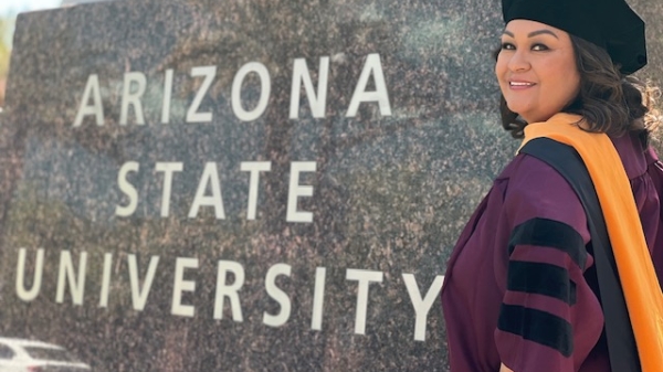Aurelia Taylor smiles at the camera in her maroon and black graduation regalia next to a sign that says Arizona State University