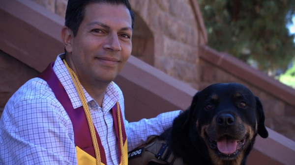 A man wearing a graduation stole sits on steps next to his dog
