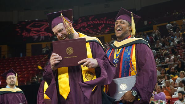 Graduates holding their degrees and wearing graduation regalia laughing.