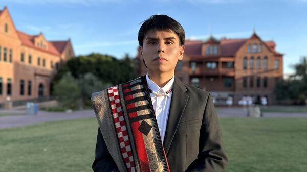 Young man wearing a suit with a Native American blanket draped over his shoulder standing in front of a university building.