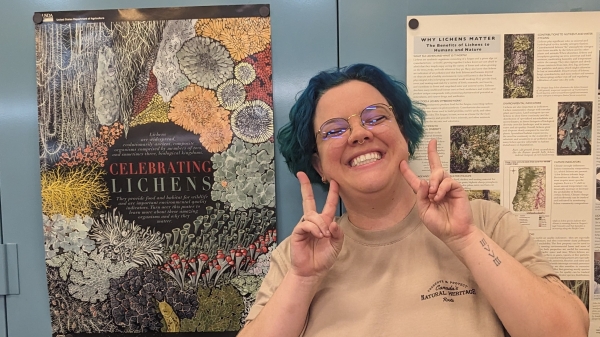 A person with blue hair smiling and holding up peace signs in front of a lichen poster.