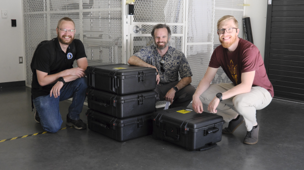 Three men kneeling next to a stack of cases and smiling.