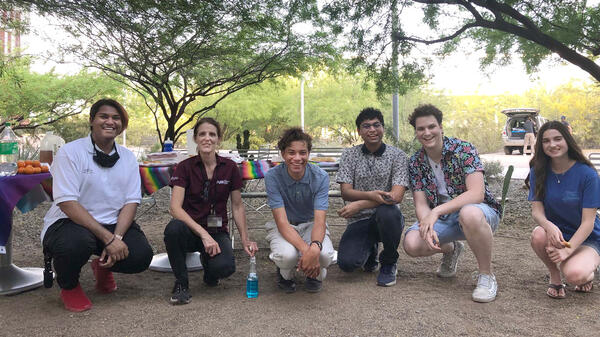 A group of smiling students poses for a photo outside under trees.