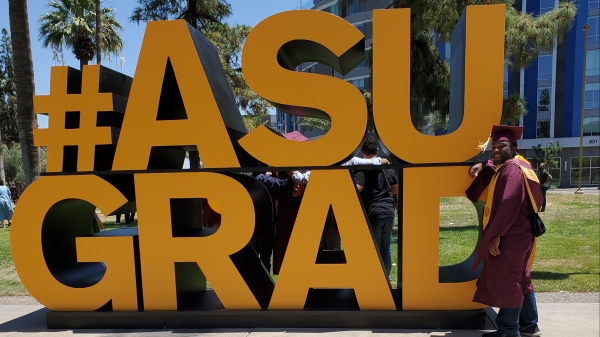 Man wearng a cap and gown standing next to a sign that reads "#ASU GRAD."