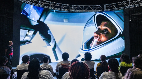 People watching presentation on large screen of woman in space helmet