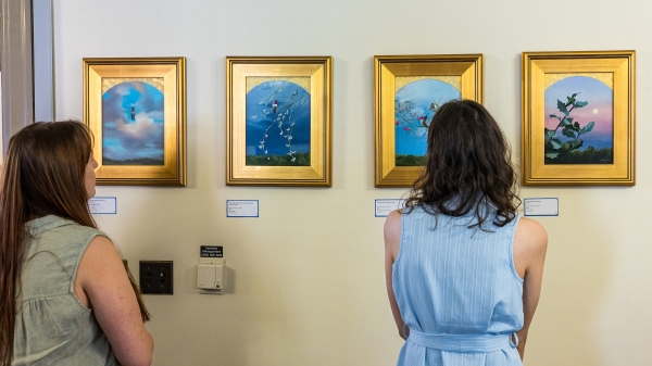 Two women examine paintings of hummingbirds hung on a wall