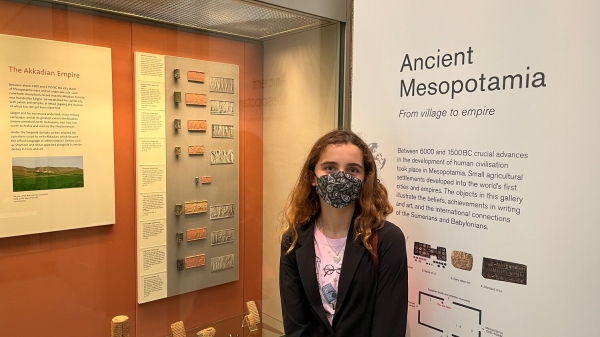 Aanya Sosa is wearing a mask and stands in front of a sign reading "Ancient Mesopotamia" 