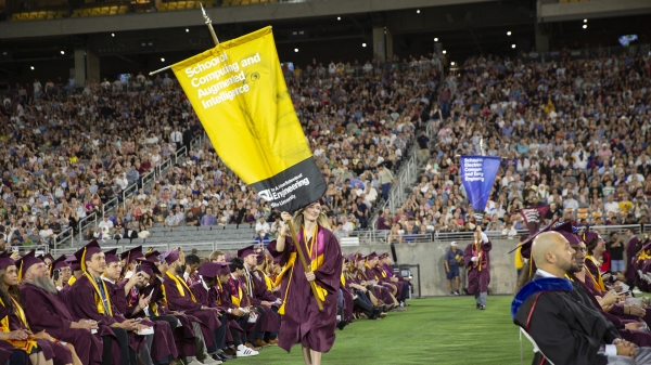 Emily Williamson carries the gonfalon for the School of Computing and Augmented Intelligence down an aisle in a crowded auditorium full of seated graduates