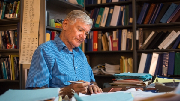 Older man wearing a blue button down shirt sits at a desk in his office with bookshelves behind him