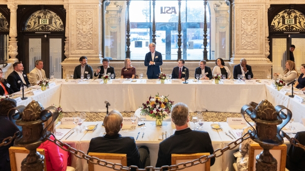 People seated at four long tables that form a rectangle for a dinner, with one person standing and talking in the middle