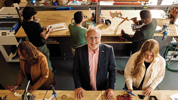 A man with glasses in a dark blazer and salmon button-down shirt smiles at the camera in the middle of a workshop full of students creating things