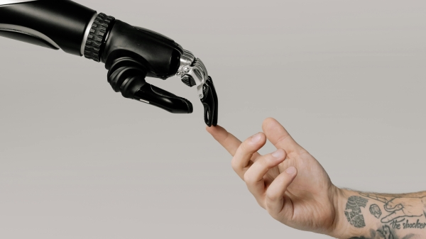 A robotic arm reaches out from the left side of the image to touch a human arm with tattoos reaching from the right of the image. The orientation is a nod to Michaelangelo's "The Creation of Adam."