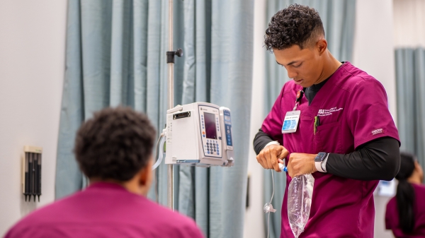 A nursing student secures a line to an IV bag in a simulation lab while a classmate looks on. Both students are in maroon scrubs