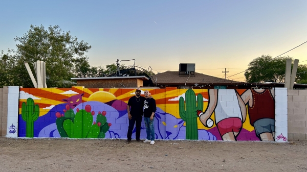 Two men stand in front of a mural of people hiking in a desert landscape.