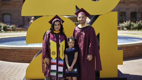 A family of three with the child standing in the middle pose in front of a large 2024 sign on ASU's Tempe campus wearing graduation attire.