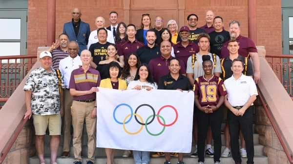 A group of people pose for a photo on steps while holding the Olympic flag