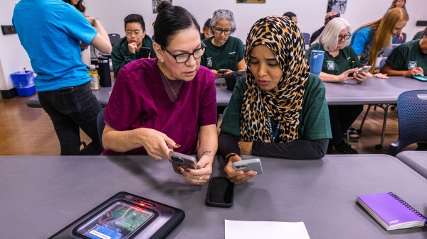 Two women sit at a table with a tablet device and cellphones.