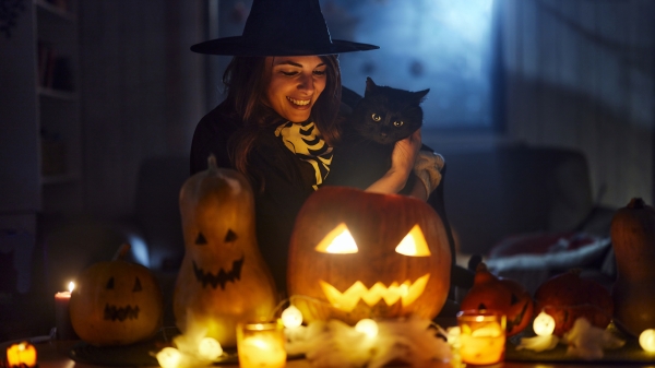 Woman dressed as a wtich holds a black cat and looks at a jack-o-lantern.