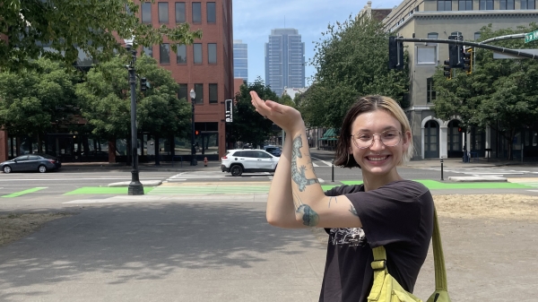 Lynae Carlin poses playfully in front of Portland city buildings with both arms "holding" a skyrise in the distance.