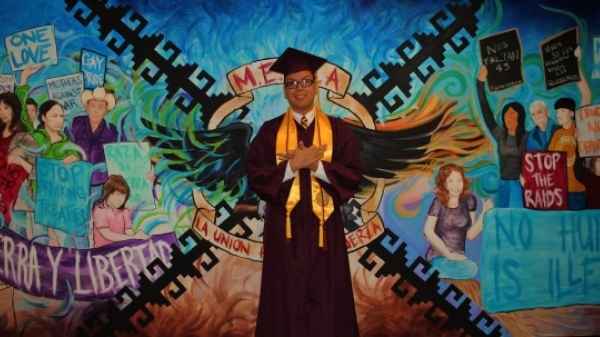 ASU alum Martine Garcia Jr. in his graduation regalia standing in front of a brightly colored mural.