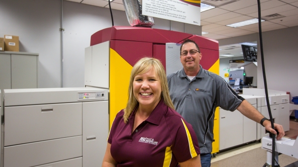 two ASU staff members posing in front of large printer