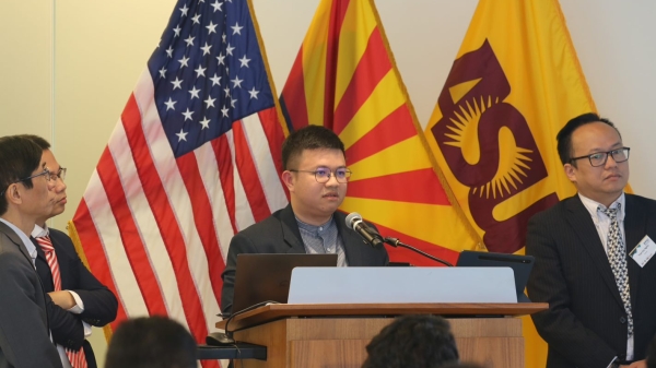 Person standing at a podium with other people surrounding them and U.S., Arizona and ASU flags behind them.