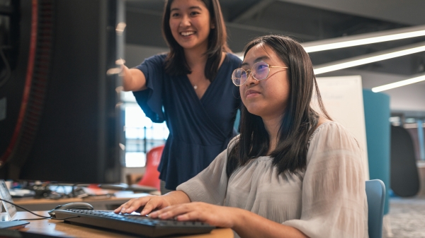 One student sits in front of a computer with another student behind them smiling.