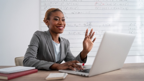 Woman smiling and waving at a laptop on a table in front of her.