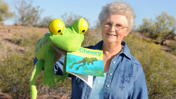 Betty Davidson holding a studded-animal frog with her children's book in its mouth, with an outdoor setting behind her.