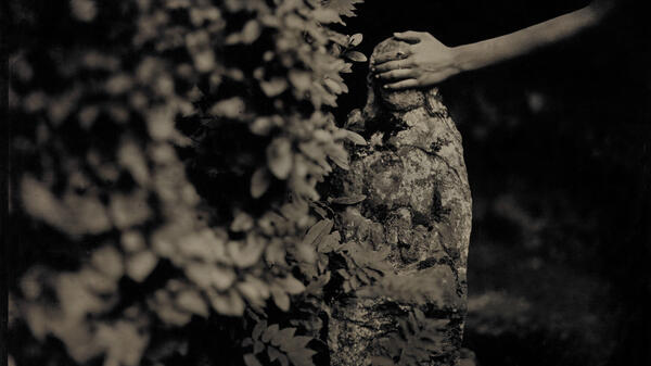 Black and white photograph of hand touching Virgin Mary statue covered in moss