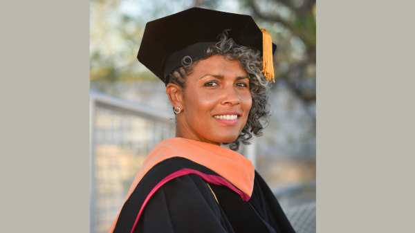 Dawn Augusta smiles at the camera. She is wearing her Doctorate Graduation Regalia complete with black hat, gold tassel and black gown with maroon trimming. 