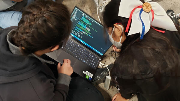 Students pictured from behind as they look at a laptop with coding on the screen.