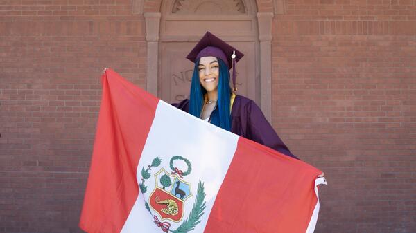 ASU alum Stefania Becerra Lavado wearing a maroon cap and gown and holding the Peru flag on the steps of ASU's Old Main building.