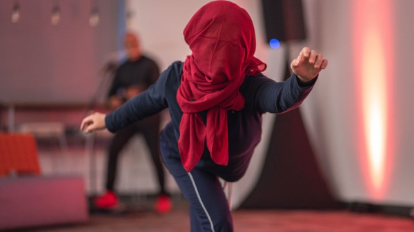 A still image of a dance performer with a red scarf covering their head. 