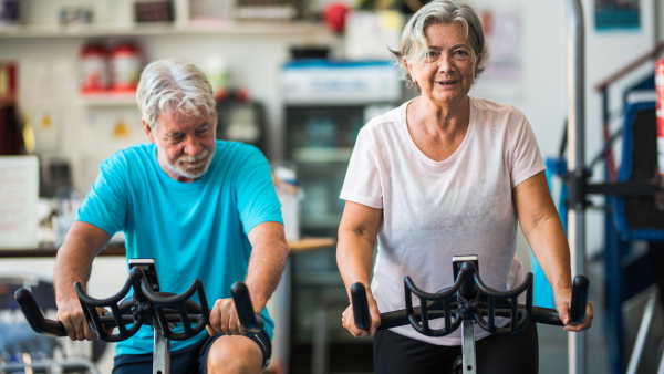 An older couple are side by side working out on stationary bikes in a gym