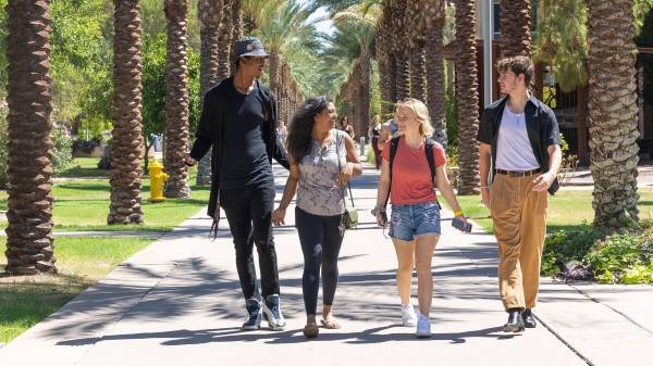 Four students smile at each other as they walk down a sidewalk lined with palm trees