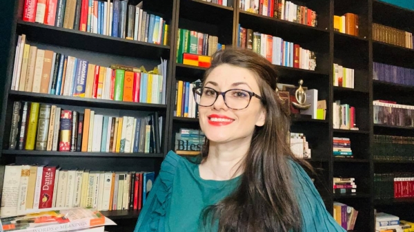 Gina Scarpete Walters smiles at the camera. She is wearing a teal blouse or dress. She has long brown wavy hair that is pulled over one shoulder. She is wearing black framed glasses and red lipstick. Behind her are many black shelves filled with books.