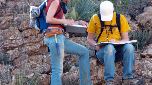 Two students in jeans and T-shirts taking notes outside in a desert environment.