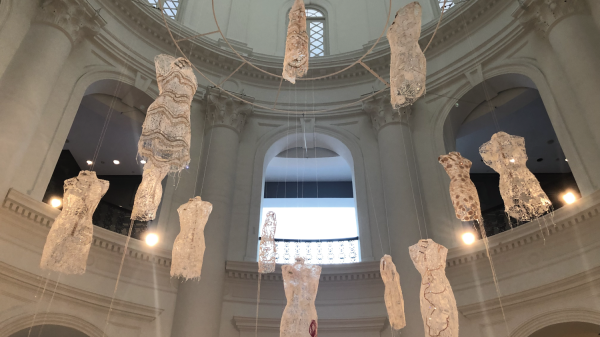 Cream-colored, brightly lit mannequins made from recycled materials hang from a large domed cathedralesque ceiling.