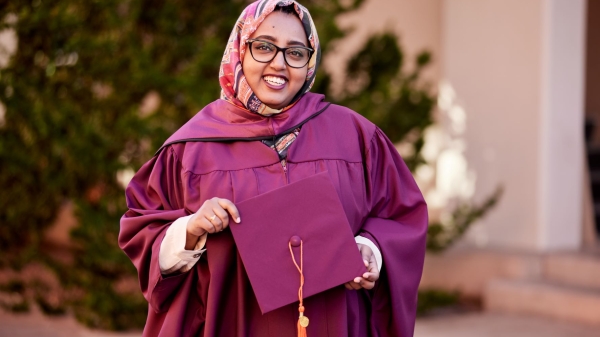 Woman in a graduation cap and gown, head scarf and glasses, smiling widely.