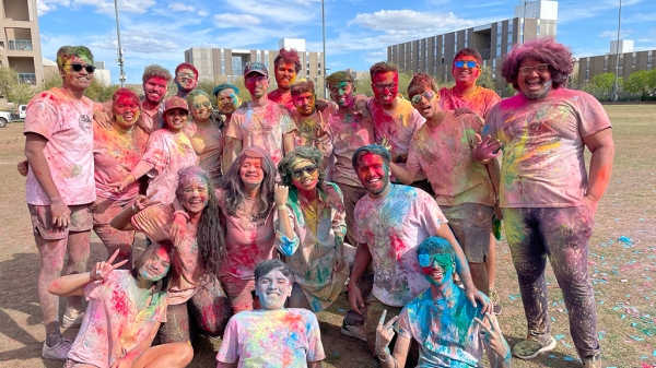 A group of people covered in colorful splashes of powder pose outside.