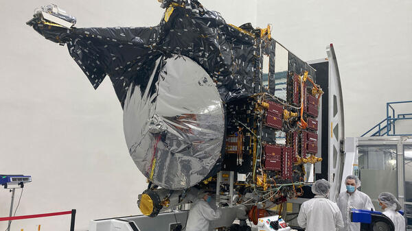 People in white lab coats and hair nets work on a spacecraft