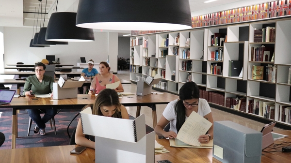 Students seated in a library at tables on which large containers are sprawled. Next to them are shelves of neatly stacked archival materials. The students are sorting through papers from the containers.