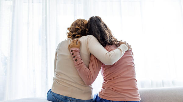 Two people seen from behind sitting next to each other, with one resting their head on the other's shoulder and their arms around each other's backs.