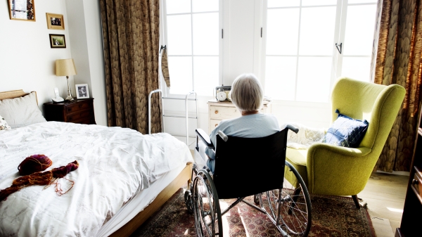 Older person seen from behind, seated in a wheelchair in a bedroom, looking out a window.