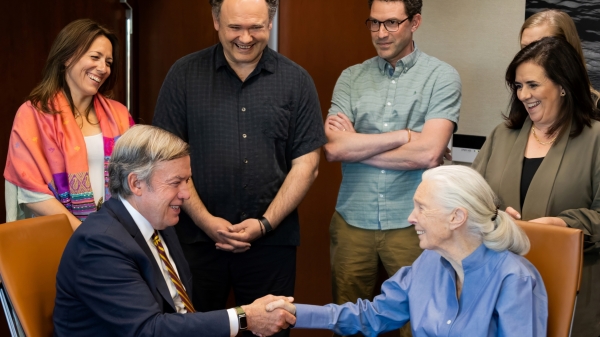 President Michael Crow shakes hands with Jane Goodall while seated at a table with five staff members standing behind them.