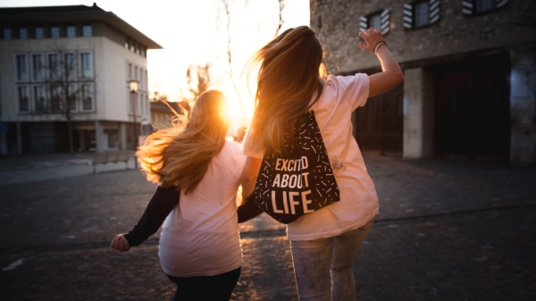 Two adolscents seen from behind skipping toward a sunset, one wearing a backpack that reads "excited about life."