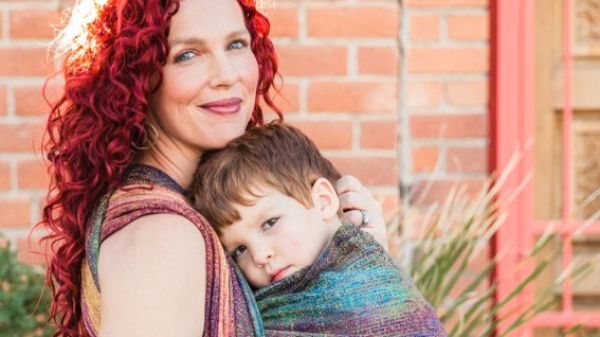Lela Rankin "babywears" her son in a colorful cloth wrap as both look at the camera, a brick building behind them.