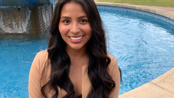 ASU grad Leslie Del Carpio smiles at the camera. She is wearing a black top, a white and black patterned skirt, and a tan blazer. She has long, wavy dark hair. Behind her is a fountain on campus.