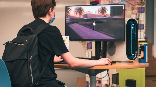Student with backpack sits at a desk with a computer monitor showing an avatar in a digital twin of the campus.